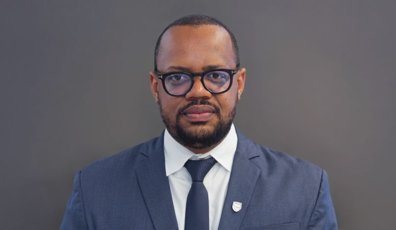 Head-and-shoulders portrait of a man in a navy suit and glasses, looking at the camera, with a gray background.