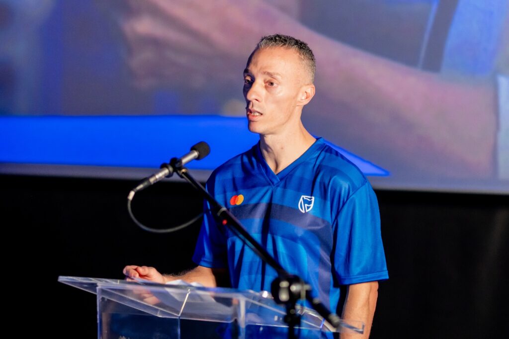 Man in a blue jersey speaks into a microphone at a clear acrylic podium on a stage.