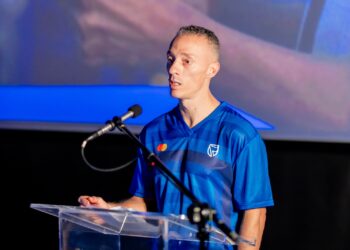 Man in a blue jersey speaks into a microphone at a clear acrylic podium on a stage.