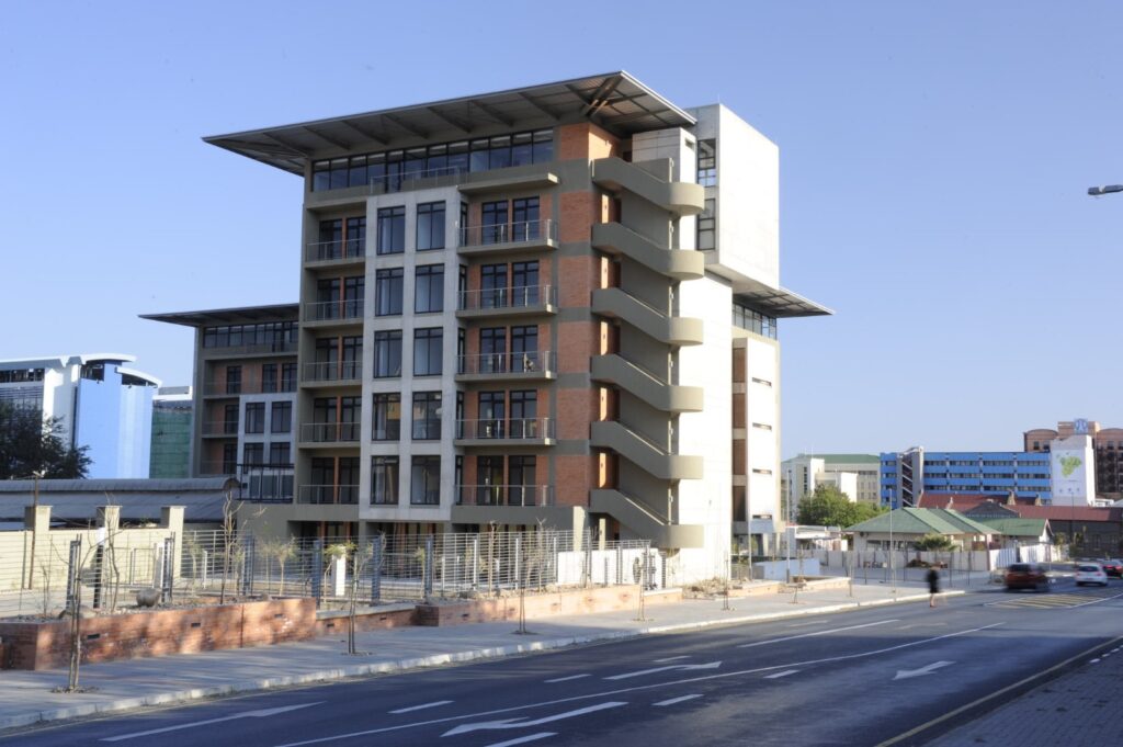 Modern six-story office building with brick and gray facade and exterior concrete staircases along a city street under a clear blue sky.