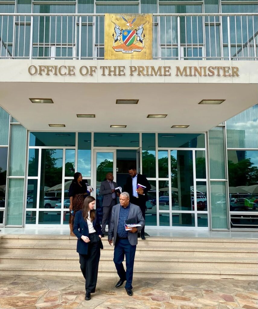 Group of professionals in business attire walking down the steps from a government building labeled 'Office of the Prime Minister' with a crest above the entrance.