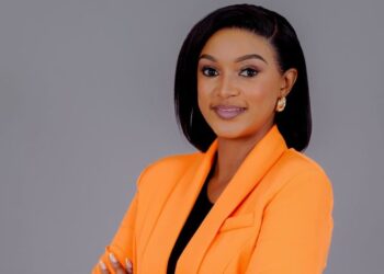Professional headshot of a woman in an orange blazer with arms crossed, smiling confidently against a gray backdrop.