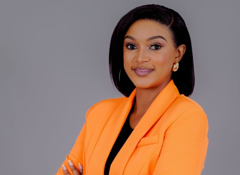 Professional headshot of a woman in an orange blazer with arms crossed, smiling confidently against a gray backdrop.