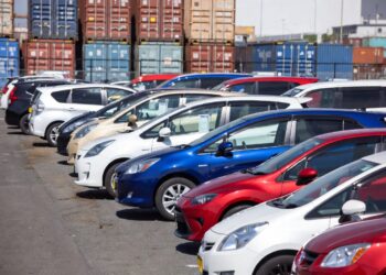 Row of colorful cars parked in a lot with stacked shipping containers in the background.