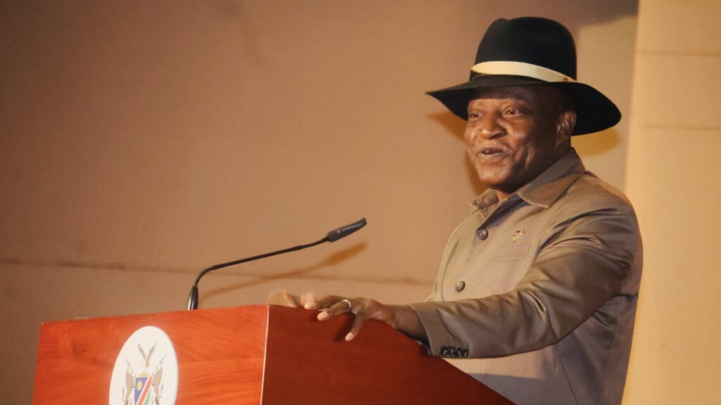 722074842_965257839681989_6726571616824513030_n - The Brief | Namibia's Leading Business & Financial News Man wearing a black hat and tan suit giving a speech at a wooden podium with a microphone in front of him.