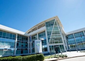 Modern glass-fronted building with a prominent central entrance, white framing, and a clear blue sky above.
