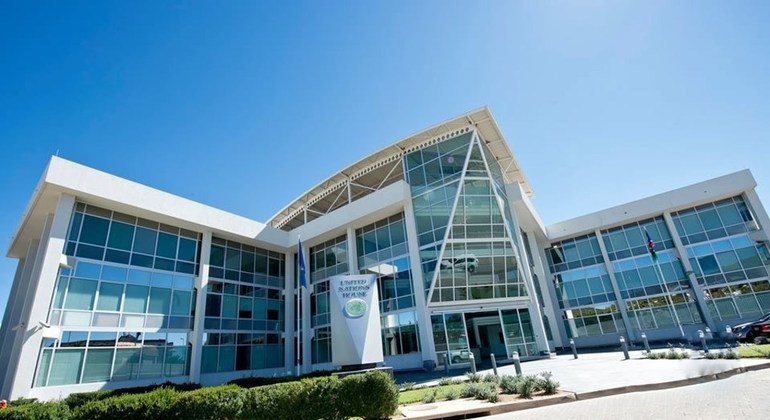Modern glass-fronted building with a prominent central entrance, white framing, and a clear blue sky above.