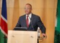 EBSON UANGUTA2 - The Brief | Namibia's Leading Business & Financial News Man in a dark suit and red tie delivers a speech at a wooden podium with flags in the background