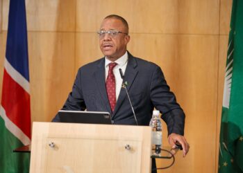 Man in a dark suit and red tie delivers a speech at a wooden podium with flags in the background