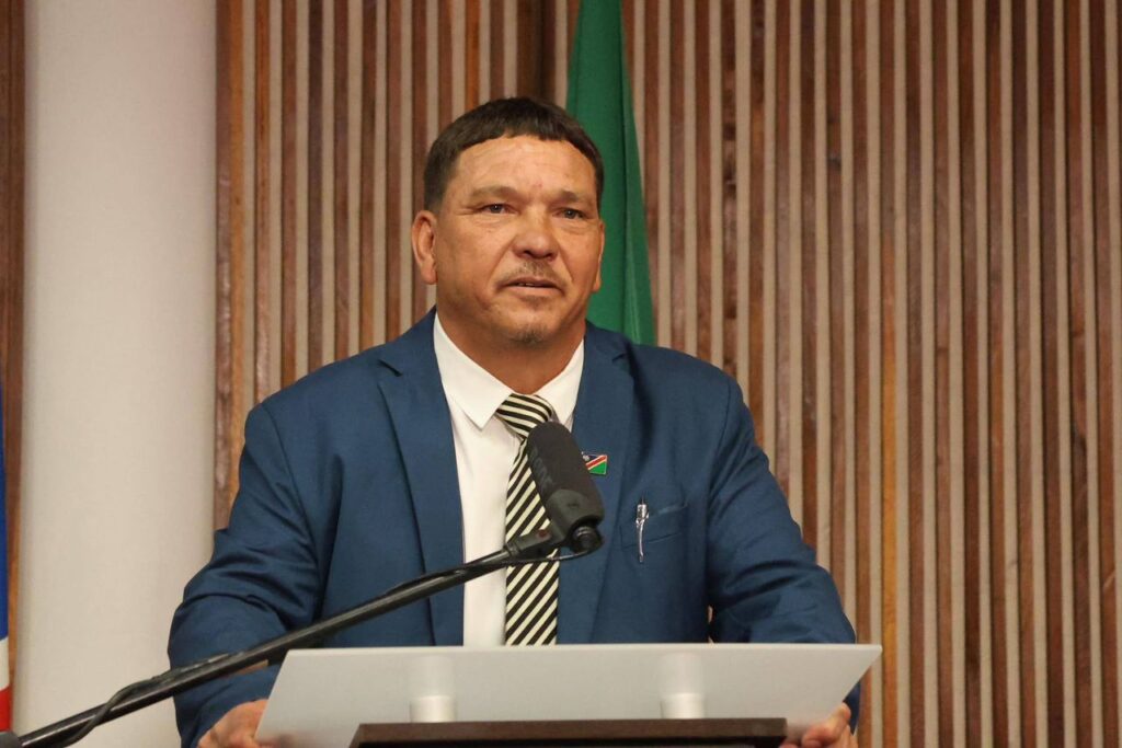Riaan Charles McNab - The Brief | Namibia's Leading Business & Financial News Man in a blue suit at a podium delivering a speech, microphone in front, wooden panel backdrop and flag behind.