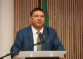 Riaan Charles McNab - The Brief | Namibia's Leading Business & Financial News Man in a blue suit at a podium delivering a speech, microphone in front, wooden panel backdrop and flag behind.