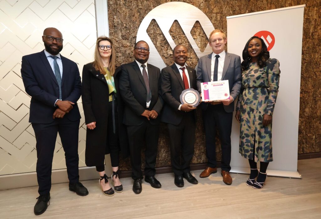 Six professionals in formal attire pose for a group photo indoors; two hold an award plaque and a certificate.