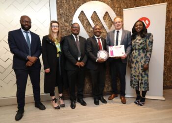 Six professionals in formal attire pose for a group photo indoors; two hold an award plaque and a certificate.