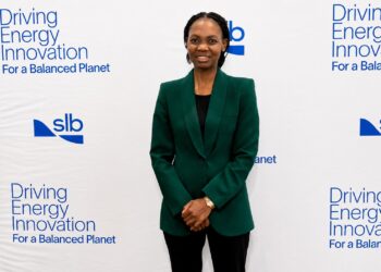 Woman in a green blazer stands in front of a white backdrop with 'Driving Energy Innovation' text and logos, hands folded.