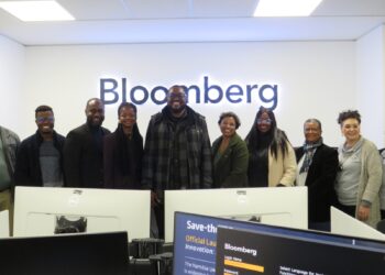 Group of coworkers posed in a Bloomberg office, smiling, with desks and computer monitors in the foreground.