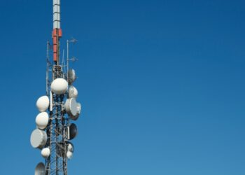 Cellular communications tower with multiple satellite dishes and antennas against a clear blue sky.