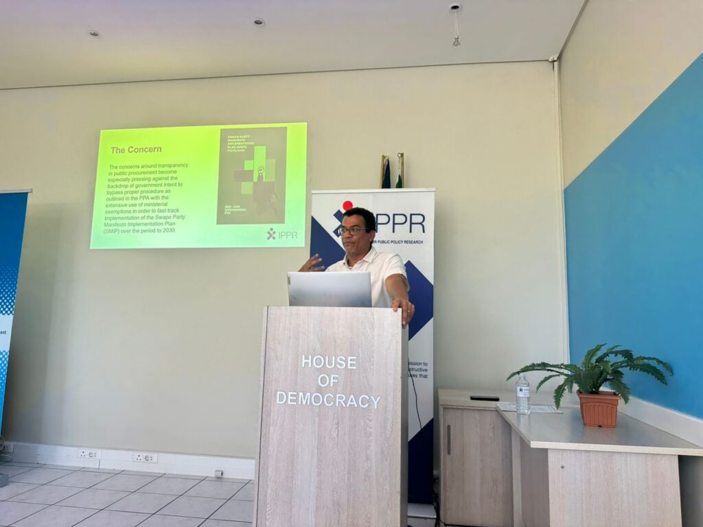 WhatsApp Image 2026-06-18 at 55252 PM - The Brief | Namibia's Leading Business & Financial News Speaker at a podium labeled 'House of Democracy' giving a talk, with a projector slide on the wall and an IPPR banner behind.