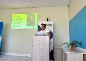 Speaker at a podium labeled 'House of Democracy' giving a talk, with a projector slide on the wall and an IPPR banner behind.