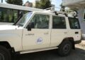 White Toyota Land Cruiser SUV with a roof rack, parked beside a building; door displays a JICA logo sticker and a small Toyota badge visible on the front panel.