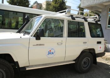 White Toyota Land Cruiser SUV with a roof rack, parked beside a building; door displays a JICA logo sticker and a small Toyota badge visible on the front panel.
