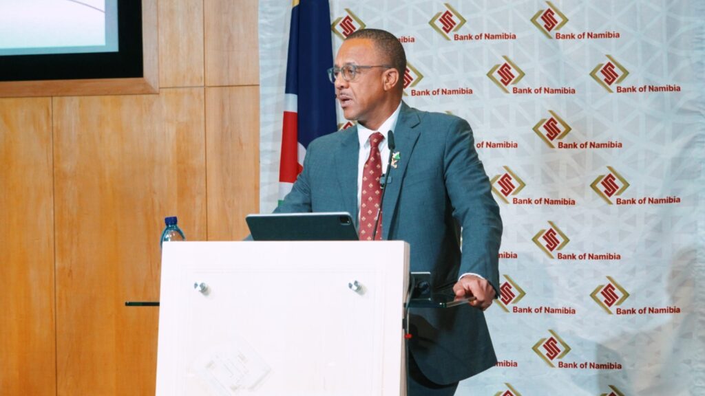 Man in teal suit delivers a presentation at a white podium with a Namibia Bank of Namibia backdrop