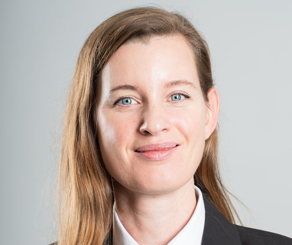 Professional headshot of a smiling woman with blue eyes and light brown hair, wearing a black blazer against a neutral background.