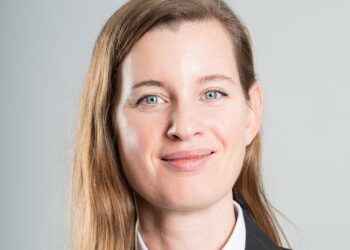 Professional headshot of a smiling woman with blue eyes and light brown hair, wearing a black blazer against a neutral background.