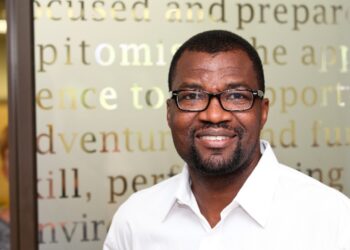 Smiling man wearing a white shirt and glasses, standing in front of a frosted glass wall with large text.