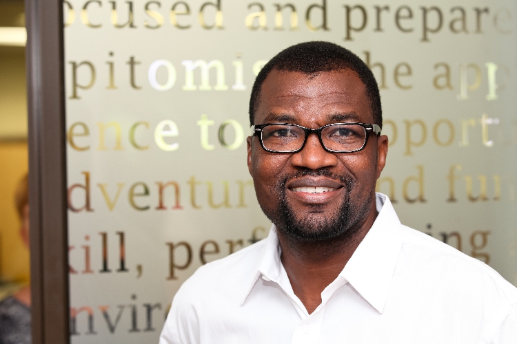 Josephat Mwatotele - The Brief | Namibia's Leading Business & Financial News Smiling man wearing a white shirt and glasses, standing in front of a frosted glass wall with large text.