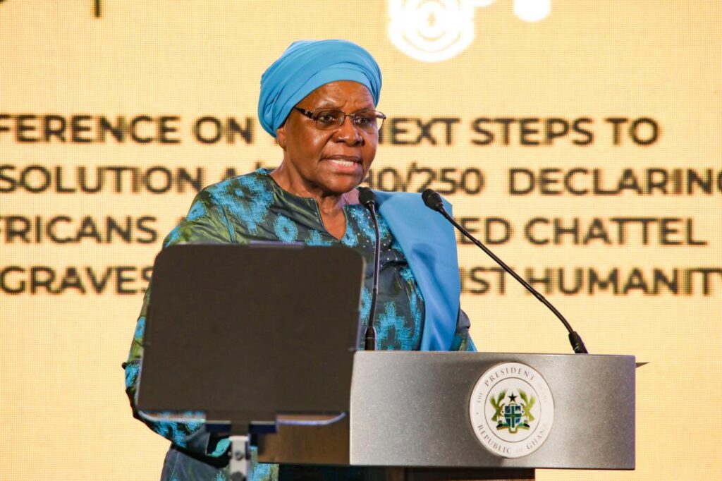 Woman in a turquoise outfit and blue headwrap delivering a speech at a podium with a presidential seal behind her.