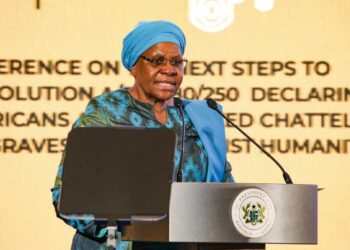 Woman in a turquoise outfit and blue headwrap delivering a speech at a podium with a presidential seal behind her.