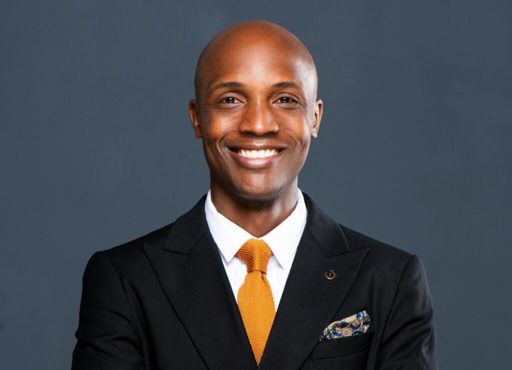 Smiling Black man in a black suit, white shirt, and orange tie against a dark gray background.