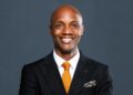 Smiling Black man in a black suit, white shirt, and orange tie against a dark gray background.