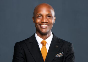 Smiling Black man in a black suit, white shirt, and orange tie against a dark gray background.