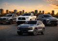 Five modern vehicles (two SUVs, a pickup, and two more SUVs) parked in a row on a paved lot with a city skyline at sunset in the background.