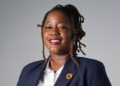 Juanita Kavandara - The Brief | Namibia's Leading Business & Financial News Portrait of a smiling Black woman in a navy blazer and white shirt, with a small lapel pin, against a gray background.",