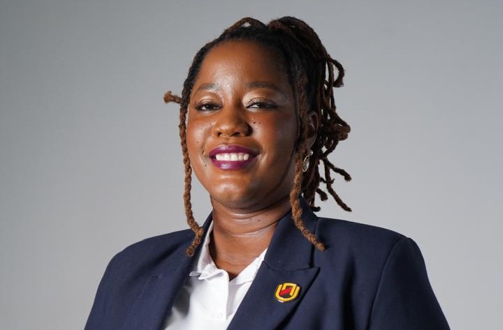 Juanita Kavandara - The Brief | Namibia's Leading Business & Financial News Portrait of a smiling Black woman in a navy blazer and white shirt, with a small lapel pin, against a gray background.",