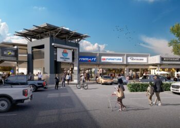 A sunny outdoor shopping plaza with a central entrance, storefronts, and people walking or cycling near parked cars.