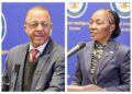 Two officials at a press briefing, a man on the left speaking into a handheld mic and a woman on the right at a podium, both in suits against a blue backdrop.