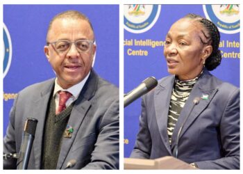 Two officials at a press briefing, a man on the left speaking into a handheld mic and a woman on the right at a podium, both in suits against a blue backdrop.