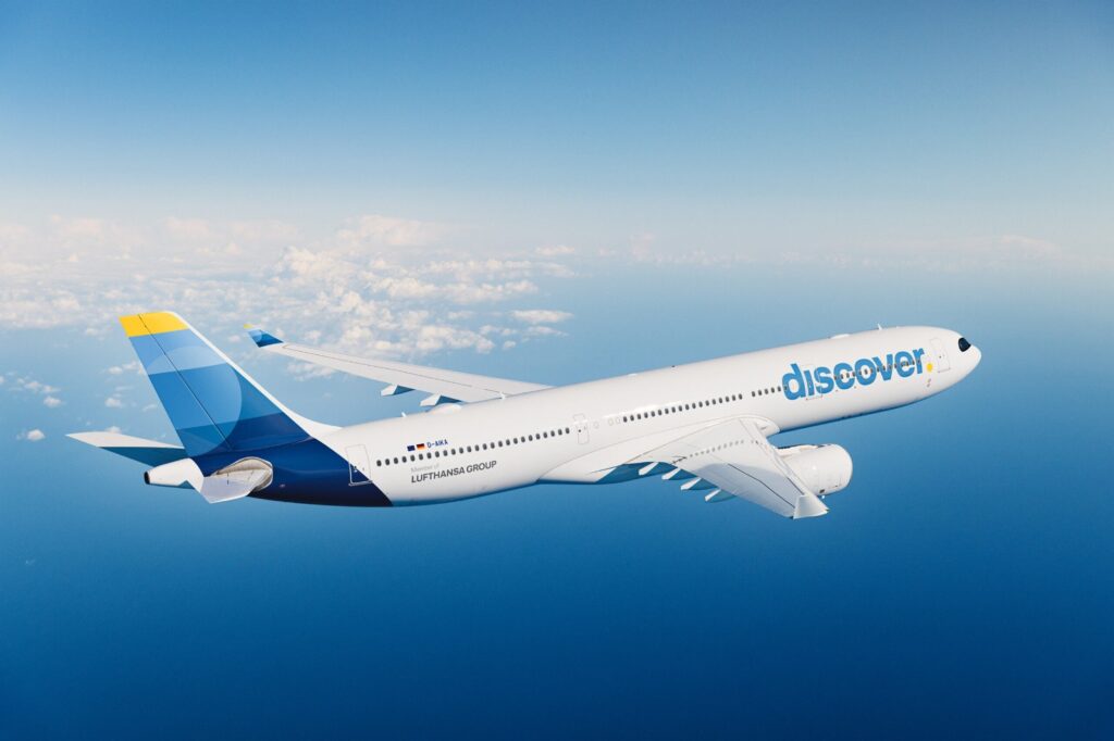 DISCOVER AIRLINE - The Brief | Namibia's Leading Business & Financial News White Discover aircraft in mid-flight over the ocean with a blue-and-yellow tail fin against a clear blue sky and scattered clouds on the horizon, showing the 'discover' logo on the fuselage.