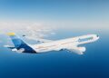 White Discover aircraft in mid-flight over the ocean with a blue-and-yellow tail fin against a clear blue sky and scattered clouds on the horizon, showing the 'discover' logo on the fuselage.