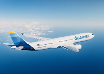 White Discover aircraft in mid-flight over the ocean with a blue-and-yellow tail fin against a clear blue sky and scattered clouds on the horizon, showing the 'discover' logo on the fuselage.