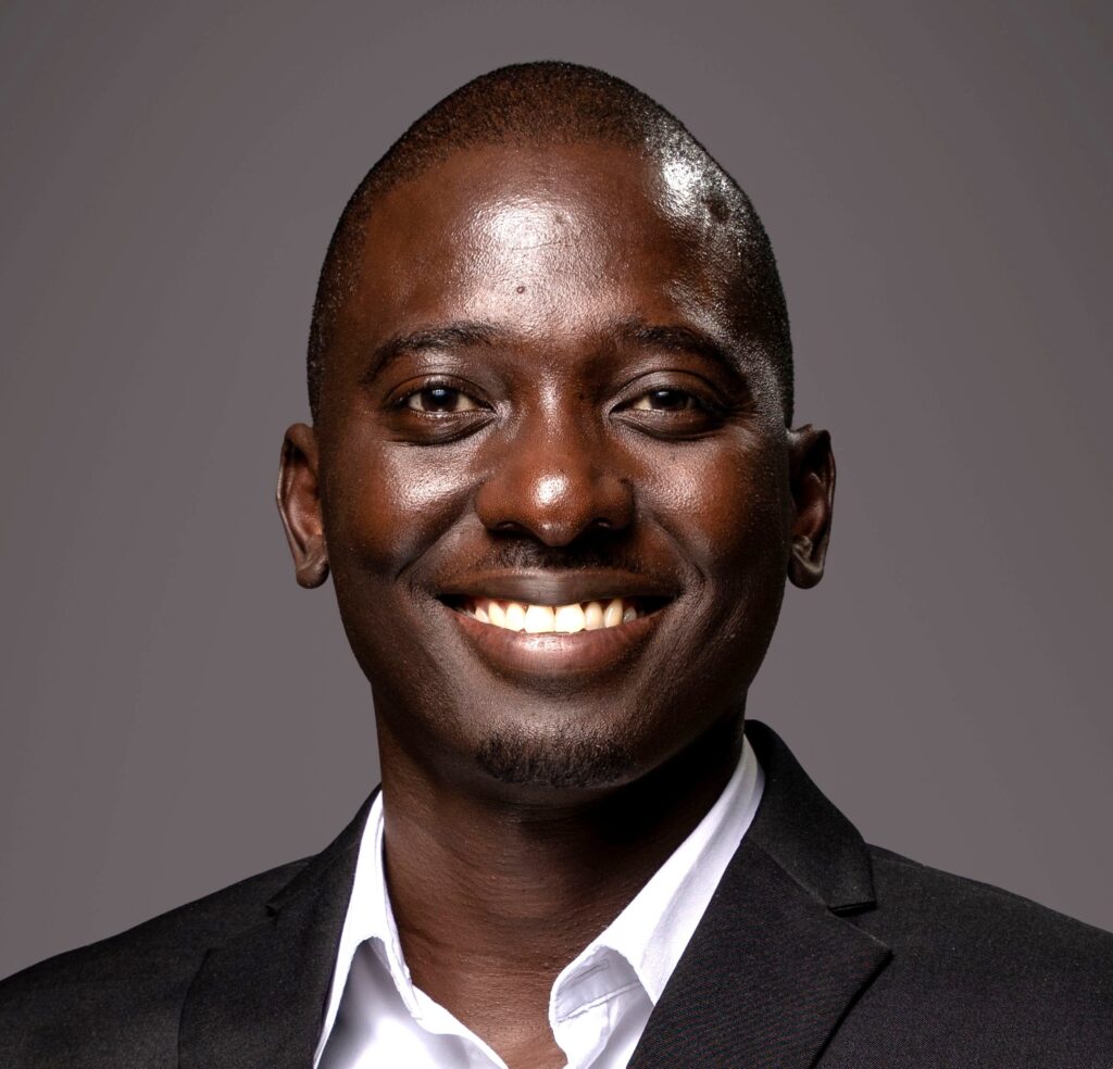Close-up portrait of a smiling Black man in a dark suit and white shirt against a gray background, showing his teeth.