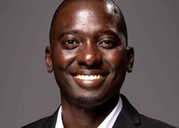 Close-up portrait of a smiling Black man in a dark suit and white shirt against a gray background, showing his teeth.