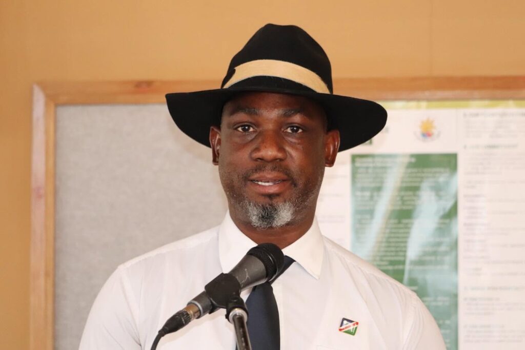 Man in a white shirt and dark tie speaks into a microphone, wearing a black fedora with a light band, indoors at a podium.