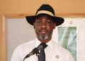Hofni Iipinge - The Brief | Namibia's Leading Business & Financial News Man in a white shirt and dark tie speaks into a microphone, wearing a black fedora with a light band, indoors at a podium.