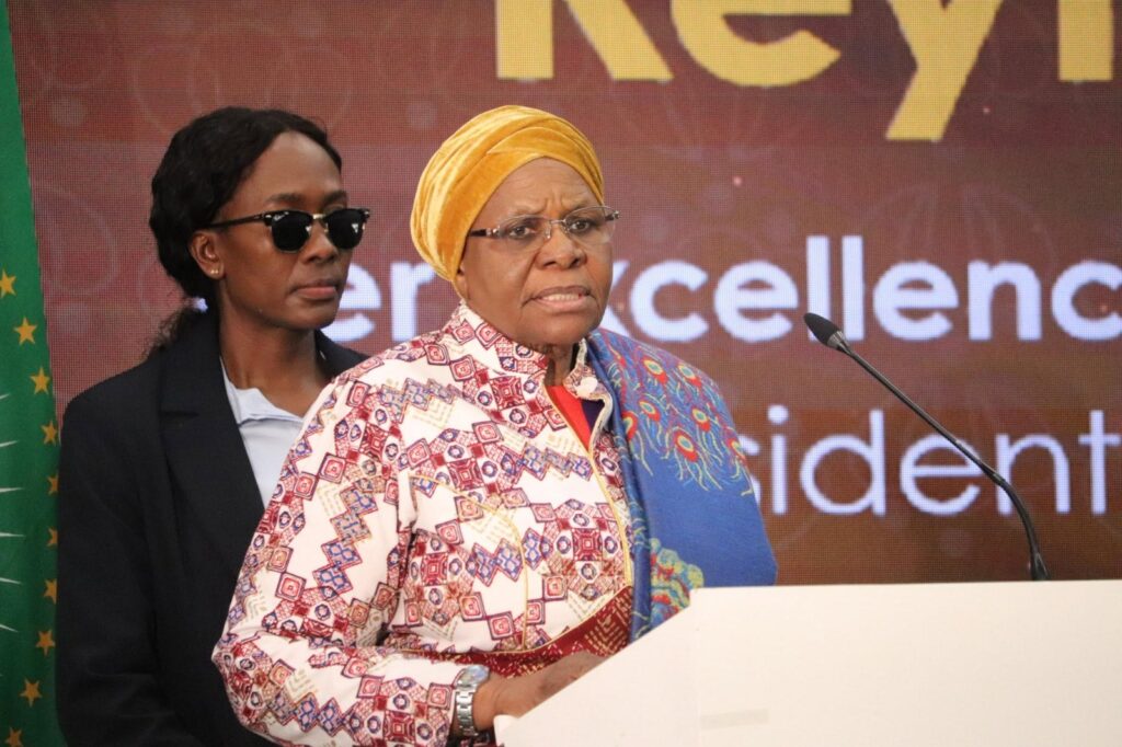 Netumbo - The Brief | Namibia's Leading Business & Financial News Woman in a colorful patterned dress and yellow headwrap speaks at a podium, with a person in sunglasses standing behind.