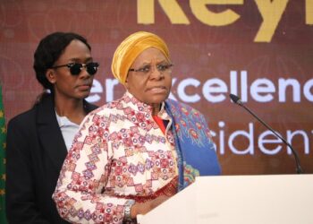 Woman in a colorful patterned dress and yellow headwrap speaks at a podium, with a person in sunglasses standing behind.