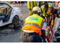 Emergency responders in high-visibility vests assist a person on a stretcher at the scene of a car crash nearby another damaged white car and debris on the road.